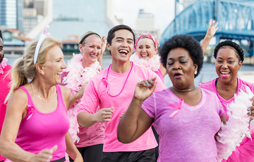 A multi-ethnic group of men and women of mixed ages wearing pink shirts, participating in a charity event to raise money for breast cancer research. They are walking together on a city waterfront, smiling and laughing.  Some are wearing pink tutus and feather boas.
