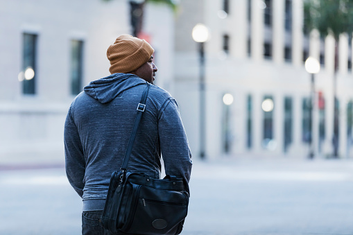 Rear view of a mature Hispanic man in his 40s walking alone on a city street. It is early morning or evening, with street lights illuminated. He is in casual clothing, wearing a hooded shirt and knit cap. He has a serious expression on his face, perhaps a little sad.