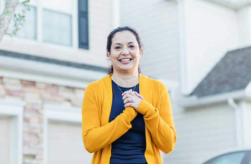 A mature Hispanic woman in her 40s standing outdoors by the driveway of her home, smiling at the camera with her hands clasped.