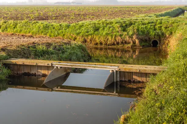 Small weir made from stainless steel and words planks and beams reflected in the mirror-smooth water of a ditch in an agricultural landscape in the Netherlands.