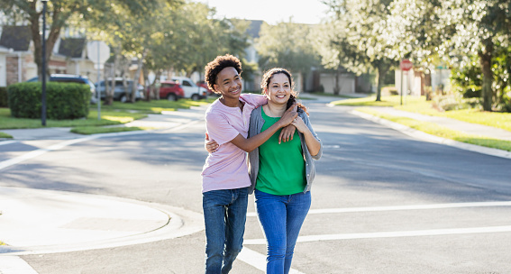 A mature Hispanic woman, in her 40s, walking with her mixed race Hispanic and African-American 14 year old teenage son outdoors along a street in their residential neighborhood. Her arm is around her the teen's waist, and his arm is draped affectionately over his shoulder.