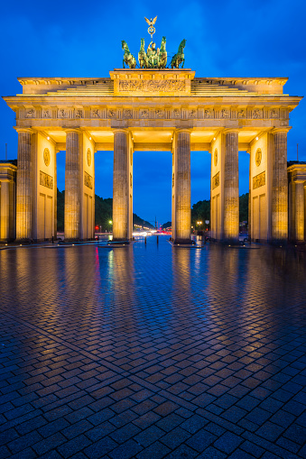 Warm spotlights illuminating the golden stonework of the Brandenburg Gate, iconic symbol of Berlin and Germany reflecting in the cobbles of Pariser Platz under blue dusk skies.