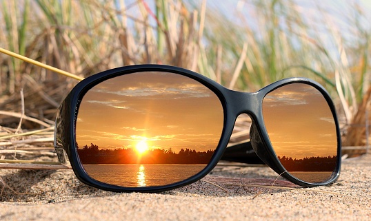 Sunset reflecting in black glass lenses laying in the sand with water and dune grasses. Peace and rest concept