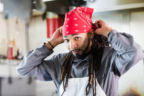 Mixed race man with dreadlocks in commercial kitchen A young mixed race African-American, Hispanic and Native American man in his 20s working as a chef in a restaurant, in the kitchen. He is wearing an apron, putting on a do rag. do rag stock pictures, royalty-free photos & images