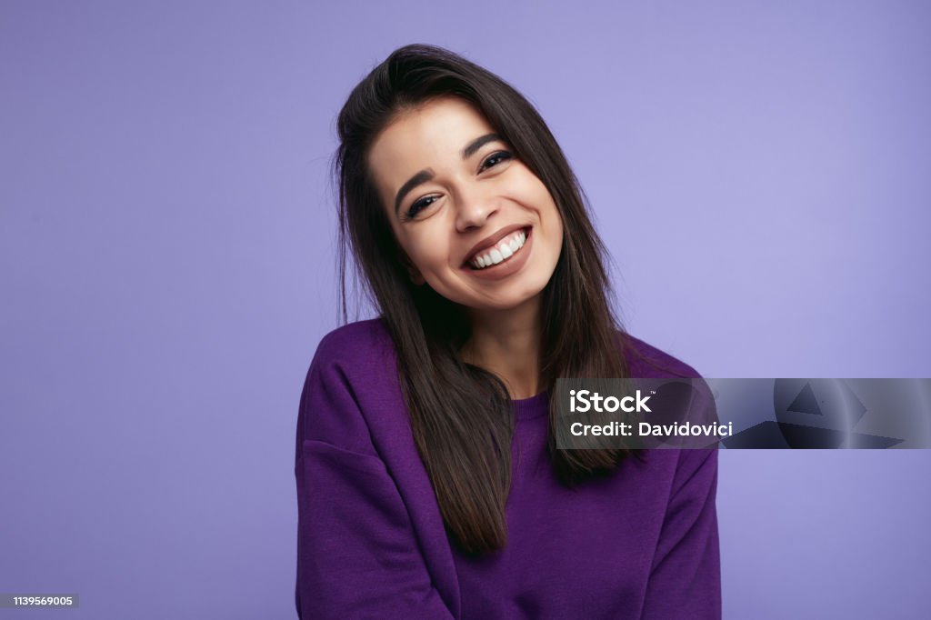 Portrait of pretty adult girl, broad smile, wears purple sweatshirt, tilts head and looks with joy, isolated over violet wall Portrait of pretty adult girl, broad smile, wears purple sweatshirt, tilts head and looks with joy, dressed casually, isolated over violet background Purple Stock Photo