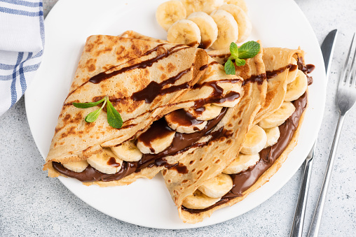 Crepes or blini stuffed with chocolate hazelnut spread, banana on a white plate. Closeup view