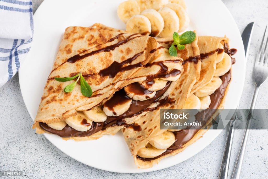 Crepes or blini stuffed with chocolate, banana Crepes or blini stuffed with chocolate hazelnut spread, banana on a white plate. Closeup view Crêpe - Pancake Stock Photo