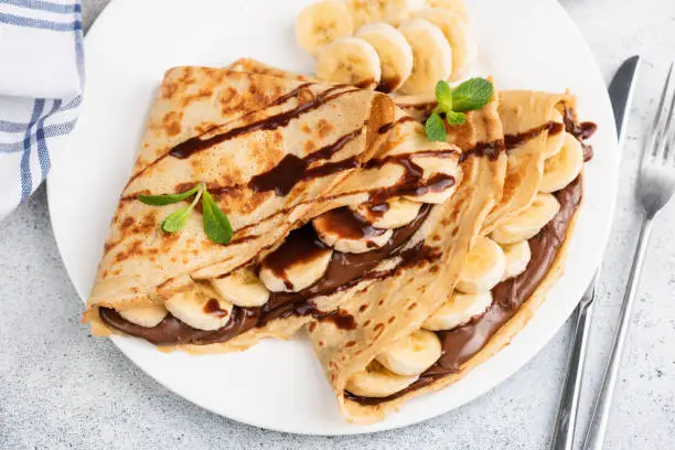 Crepes or blini stuffed with chocolate hazelnut spread, banana on a white plate. Closeup view