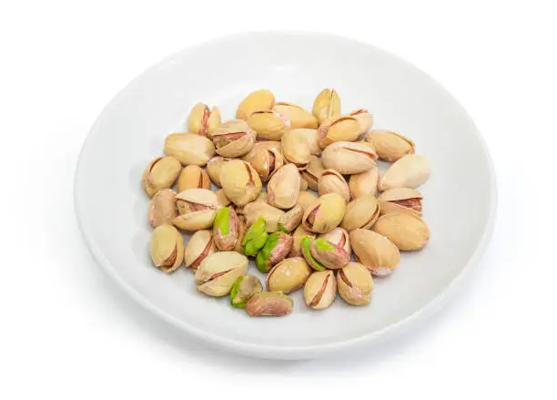 Roasted salted pistachio nuts peeled from shells and nuts with partly open shells on the white saucer on a white background