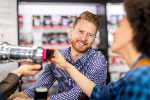 female shopper jokingly putting a small compact camera on a large silver telescopic lens held by her partner in a camera equipment store - retail occupation flash imagens e fotografias de stock