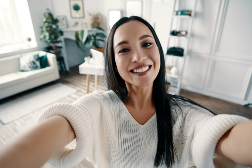 Attractive young woman looking at camera and smiling while taking selfie at home