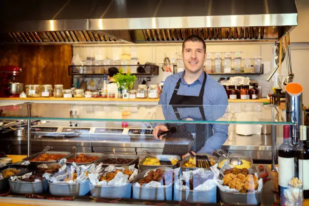 Photo of Portrait of smiling owner standing at his eatery serving food behind counter, young entrepreneur wearing apron running small restaurant, happy man working