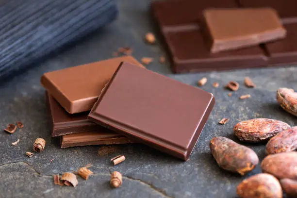 Four squares of dark and milk chocolate with small chocolate bits on grey slate next to cocoa beans. Blurred chocolate in the  background.