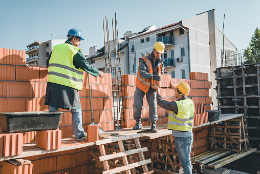 Young Building Workers is Helping out his Experienced Colleague at the Roof of the Construction Site. Teamwork by Three Builders with Protective Helmets who are Building a Wall of Bricks at the Building Site.
