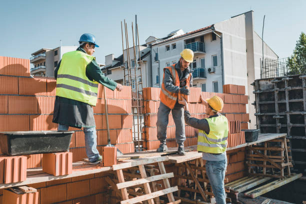 Construction Workers are Building a Brick Wall at the Rooftop of the Building Young Building Workers is Helping out his Experienced Colleague at the Roof of the Construction Site. Teamwork by Three Builders with Protective Helmets who are Building a Wall of Bricks at the Building Site. bricklayer stock pictures, royalty-free photos & images