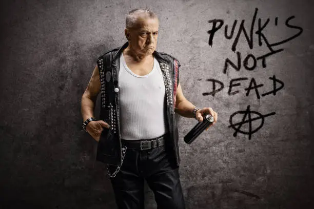 Photo of Mature male punker holding a wall spray and leaning against a rusty gray wall with written punk's not dead and anarchy sign