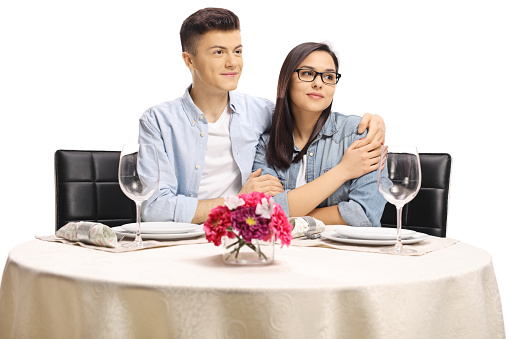 Teenage male and female in an embrace sitting at a restaurant table looking away isolated on white background