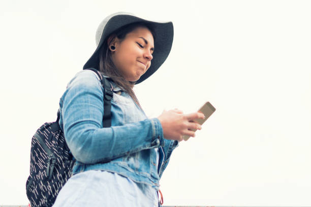 pretty hispanic girl smiling and chatting on her phone, city break, solo traveler, social media, one young woman tourist using smartphone, enjoyment, sunset light, portrait, back lit, travel, exploration, tourism, outdoors, generation z, springtime - back lit women one person spring imagens e fotografias de stock