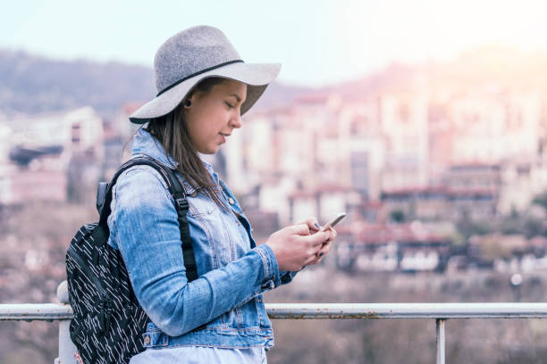 pretty hispanic girl smiling and chatting on her phone, city break, solo traveler, social media, one young woman tourist using smartphone, enjoyment, sunset light, portrait, back lit, travel, exploration, tourism, outdoors, generation z, springtime - back lit women one person spring imagens e fotografias de stock