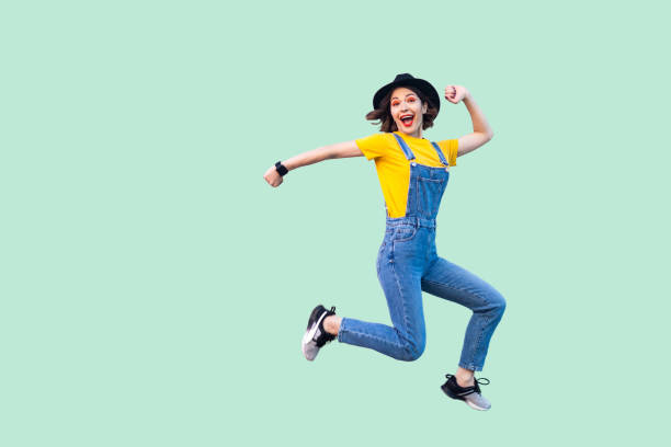 파란색 데님 바지, 노란색 셔츠와 검은 모자 점프와 카메라를 보고에서 행복 깜짝 예쁜 젊은 소식통 여자의 초상화를 즐길 수 있습니다. - women teenage girls jumping dancing 뉴스 사진 이미지