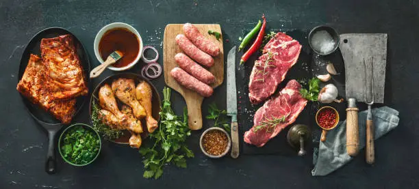 Various kinds of grill and bbq meats with vintage kitchen and butcher utensils. Chicken legs, steaks, sausages, pork ribs with herbs, spices, sauces and ingredients for grilling