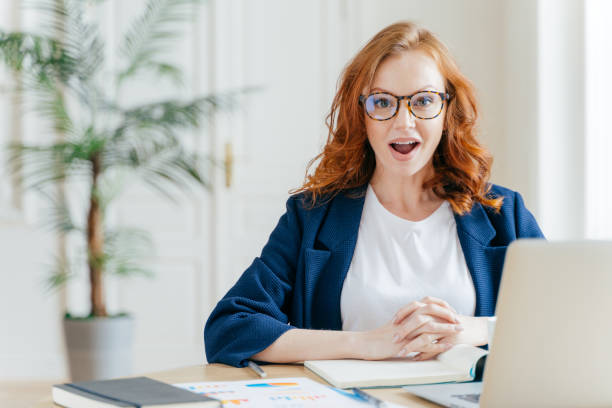 istock Surprised ginger female employee has job, develops new business srategy, poses in front of opened laptop computer, poses in workspace against office interior. Woman software developer at work. 1139704020