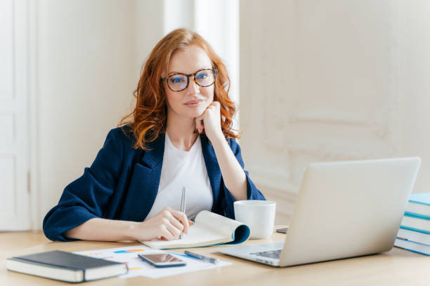 istock Redhead curly woman office worker analyzes data, makes accounting report, poses in coworking office, improves online sales, drinks coffee, wears spectacles and formal wear. Female employee indoor 1139704036