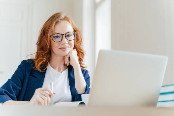 Horizontal shot of pleasant looking successful professional female lawyer learns clients case, works on modern laptop computer, dressed in formal apparel and transparent glasses, poses in office Horizontal shot of pleasant looking successful professional female lawyer learns clients case, works on modern laptop computer, dressed in formal apparel and transparent glasses, poses in office redhead stock pictures, royalty-free photos & images