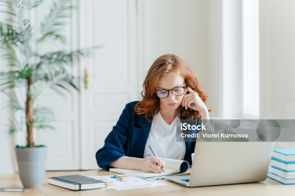 Serious confident female entrepreneur in formal clothes writes down information while watches business webinar, notes main ideas, sits at work place in front of opened computer makes notes for startup Working Stock Photo