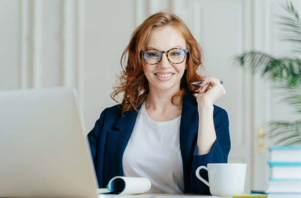 istock Glad redhead curly woman works freelance, uses laptop computer, writes down notes in notepad, drinks coffee, wears optical glasses, being in good mood, develops business company. Work concept 1139704373