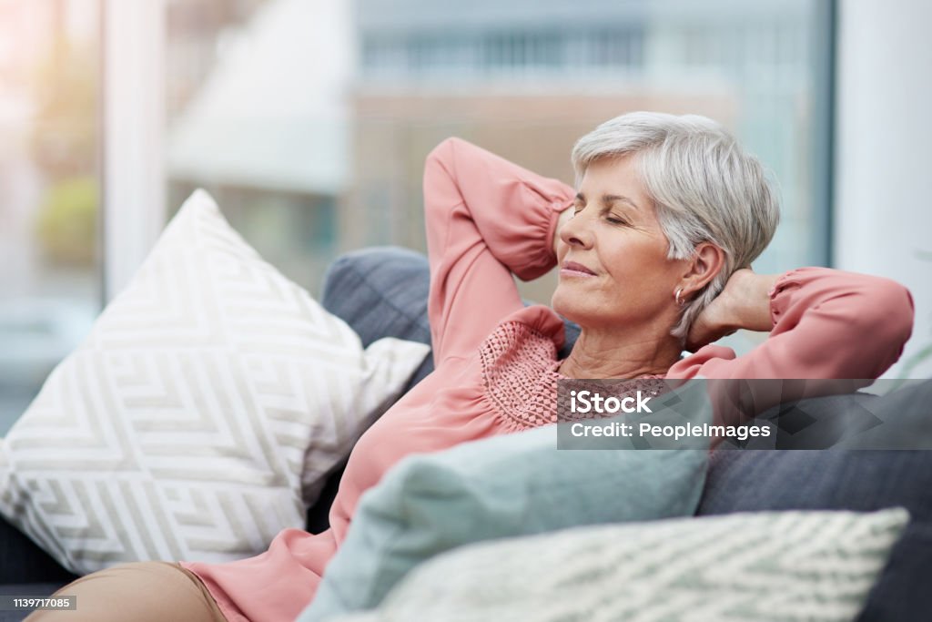 Time to kick back and relax Shot of cheerful mature woman laid back and relaxing on her sofa at home Relaxation Stock Photo