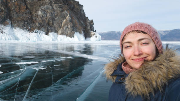 Woman traveler makes a selfie against the rock and Baikal ice. Smiling traveler in winter clothes on the background of the winter lake Baikal. stock photo