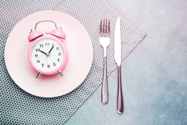 Alarm clock and plate with cutlery. Concept of intermittent fasting, lunchtime, diet and weight loss Alarm clock and plate with cutlery. Concept of intermittent fasting, lunchtime, diet and weight loss fasting stock pictures, royalty-free photos & images