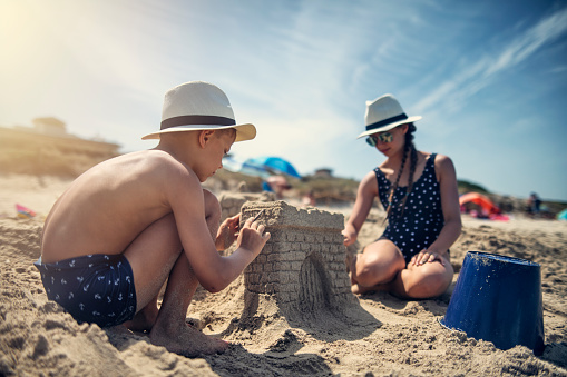 Children having fun building sandcastles on beach. Little boy is carving a sand gate to the castle. His siblings are building in the background.\nNikon D850