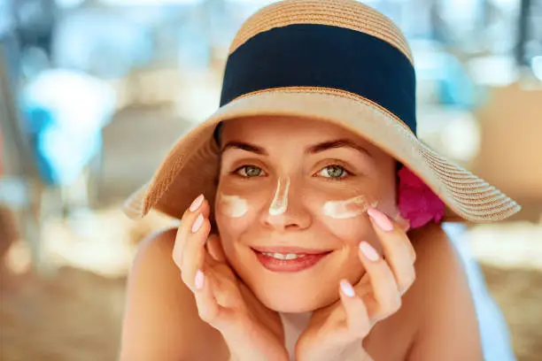 Photo of Skincare. Beauty Concept. Young pretty woman applying sun cream  and touch own face. Female in hat smear  sunscreen lotion on skin. Skin Protection and dermatology.