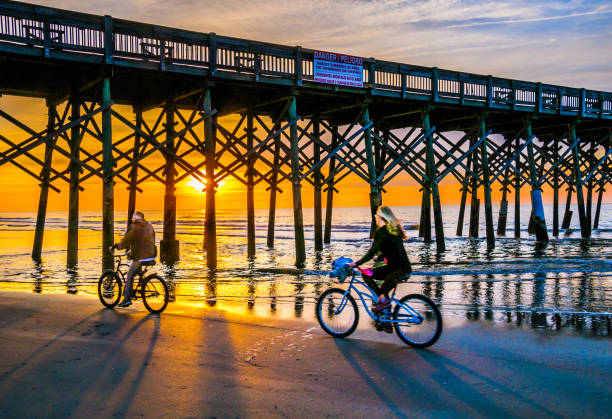 Biking on the Beach Folly Beach, South Carolina, USA -March 24, 2019-A mature couple ride their bicycles along the water's edge beneath the wooden pilings of the Folly Beach Pier as the morning sun slowly rises in the East. charleston south carolina photos stock pictures, royalty-free photos & images