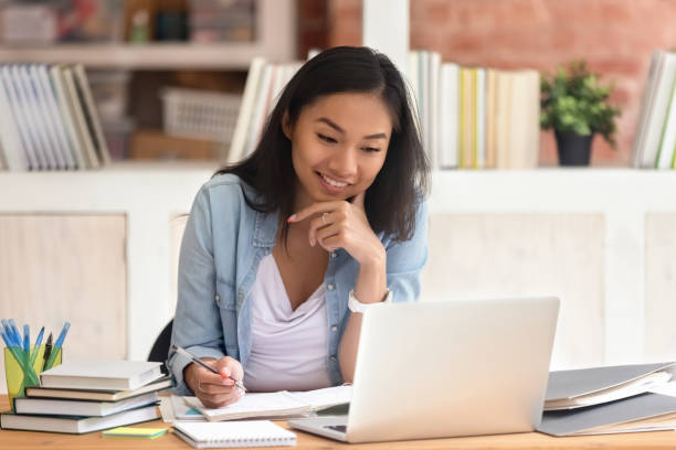 sorridente studentessa asiatica studia in biblioteca con libri per laptop - student teenager college student university foto e immagini stock