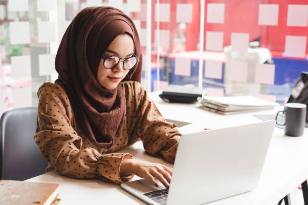 junge asiatische muslimische geschäftsfrau in schicker casual wear, die mit laptop-computer arbeitet, während sie im kreativen café sitzen. - east asian ethnicity stock-fotos und bilder