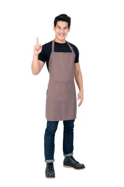 Isolated portrait of a young attractive male asian barista in brown apron smiling and  pointing hand upward