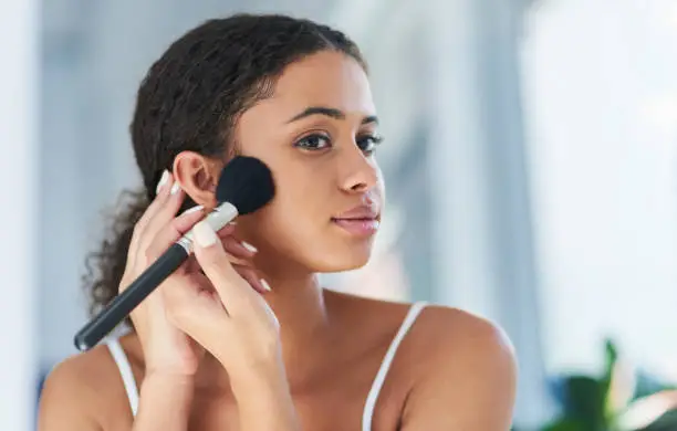 Cropped shot of a young woman applying makeup to her cheeks in the bathroom at home