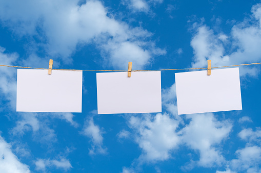 Blank photo paper hanging on a clothesline over clouds in the blue sky background