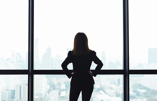 Asian business woman with arms akimbo looking out the window at city view background