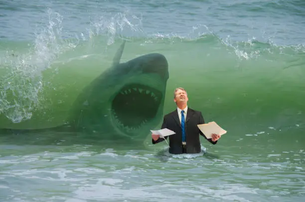 Businessman holding paperwork getting ready to be hit by a big wave and attacked by shark representing drowning in business pressure, deadlines, work stress, overworked, problems and problem solving.