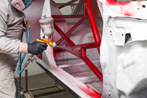 保護オーバーオールとマスクの男は、彼の手にスプレーボトルを保持し、車両修復ワークショップでの修理中に事故の後、車体のフレーム上に赤いペンキをスプレー - priming paint ストックフォトと画像