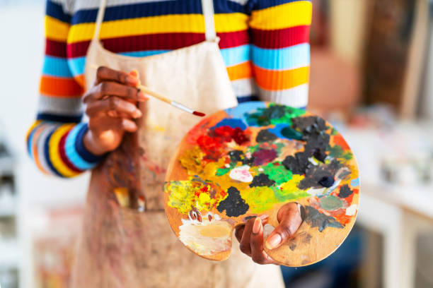 smiling young afro woman holding color palette and paintbrush - women artist painting easel imagens e fotografias de stock