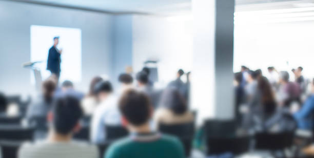 Blurred De-focused Audience in Conference Lecture Presentation Room. Corporate Presentations in Conference Hall. Seminar Speaker Giving Training to New Employees. Blurred  Hip Presenter wearing Hat. stock photo