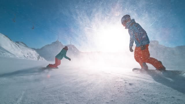 SLO MO TS Male and female snowboarder riding down a sunny slope and snow particles flying in the air