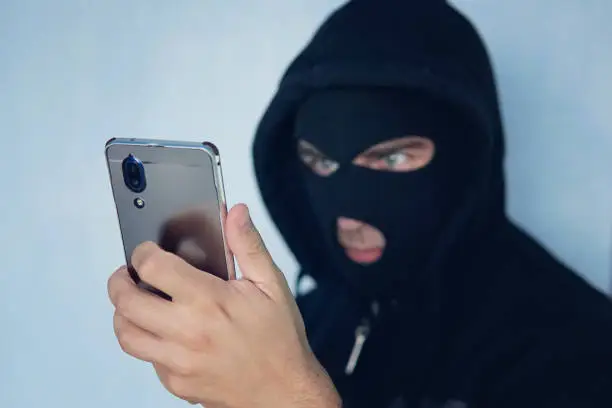 Photo of Young adult in black clothes with hidden face looks at smartphone screen. Ill-intended fraudster uses mobile. Fraudster calls. Mobile racket. Hacker hijacks by phone. Cellphone account fraud. Scam.