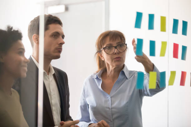 Middle aged businesswoman explaining project strategy on sticky notes Middle aged businesswoman mentor coach leader explaining project strategy showing post it sticky notes on glass wall, serious business team brainstorm work plan at corporate office stand up meeting delegating stock pictures, royalty-free photos & images