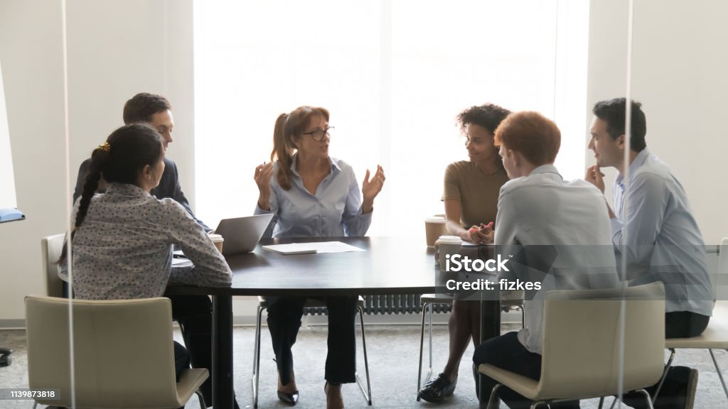 Middle-aged businesswoman speaking at diverse group negotiations at conference table Middle aged businesswoman speaking at diverse group negotiations discussing work with colleagues, female executive talking to coworkers at corporate briefing sitting at conference table in boardroom Meeting Stock Photo